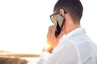 Man outdoors in Morocco talking on smartphone, wearing sunglasses and white shirt, backlit by sunset.